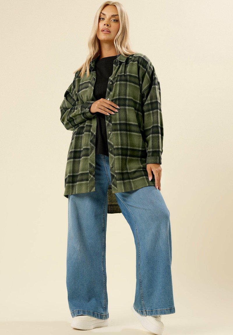 Camicia di flanella a quadri verdi con accenti neri, chiusura a bottoni, abbinata a jeans in denim blu larghi e sneakers bianche. Texture liscia.