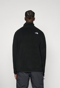 Musta fleece-takki korkealla kauluksella ja The North Face -logolla yläselässä. Pehmeä rakenne, rento istuvuus ja pitkät hihat.