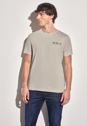 T-shirt à col rond gris clair, à manches courtes, avec une impression de texte noir sur la poitrine gauche. Porté avec un jean en denim bleu foncé.
