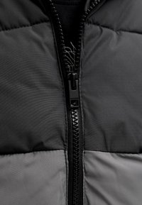 Szara kurtka typu puffer z gładkim czarnym zamkiem błyskawicznym, dwukolorowym wzorem z ciemnoszarymi i jasno szarymi sekcjami oraz miękką fakturą tkaniny.