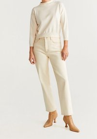 Beiger Strickpullover mit hohem Kragen kombiniert mit cremefarbenen geraden Jeans und beigen Absatz-Stiefeletten, vor einem neutralen Hintergrund präsentiert.