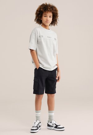 Jaunas žmogus su garbanotais plaukais, dėvintis šviesiai pilką marškinėlį su grafiniu piešiniu, juodus cargo šortus, pilkas dryžuotas kojines ir juodai baltus sportbačius, stovintis.