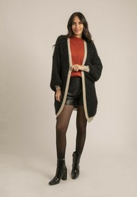 Cardigan noir en peluche avec bord beige, porté sur un pull rouille et un short en cuir, associé à des bottes à talons noires et des collants semi-transparents.