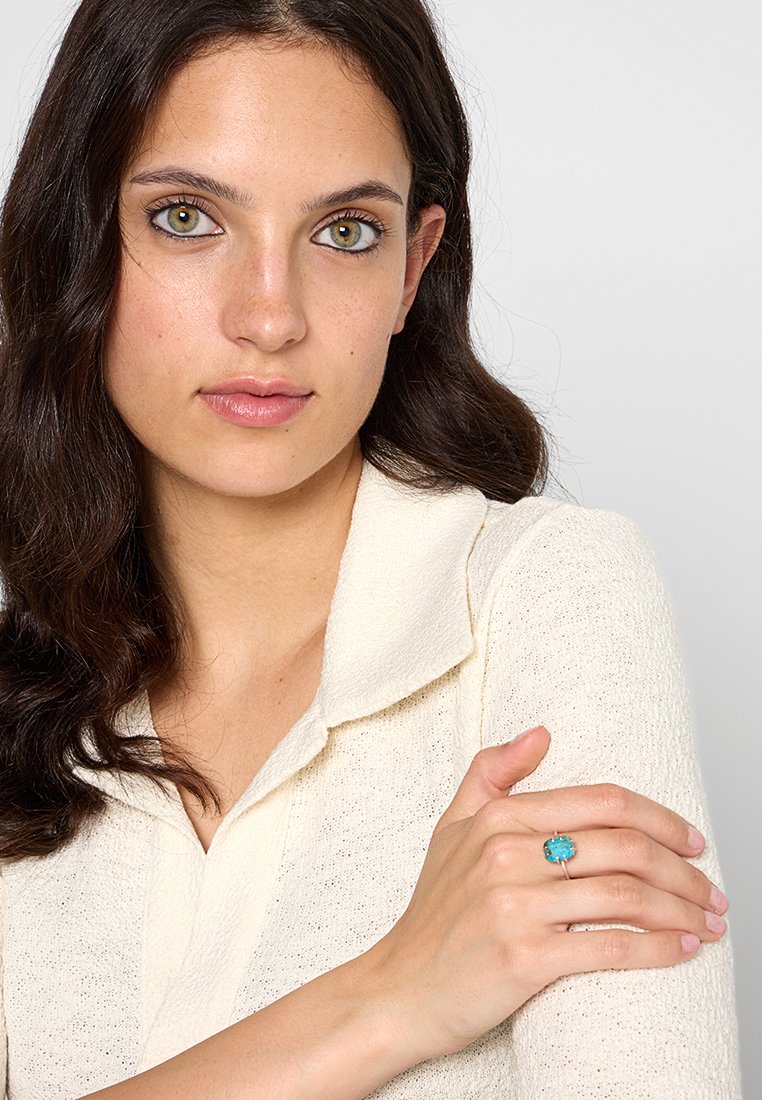 Anillo turquesa con una piedra rectangular, engastado en plata. El modelo lleva una blusa de color blanco roto con textura y cuello, con los brazos cruzados.