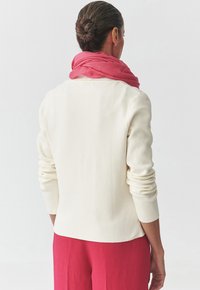 Maglione color crema con maniche lunghe, caratterizzato da una vestibilità morbida. Una sciarpa rosa leggera si drappeggia attorno al collo, aggiungendo texture.