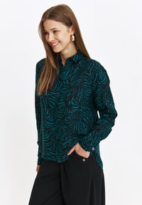 Czarna bluzka z turkusowym, abstrakcyjnym wzorem fali, długie rękawy, kołnierz zapinany na guziki, detal kieszeni oraz lekki połysk. Gładka tekstura materiału.