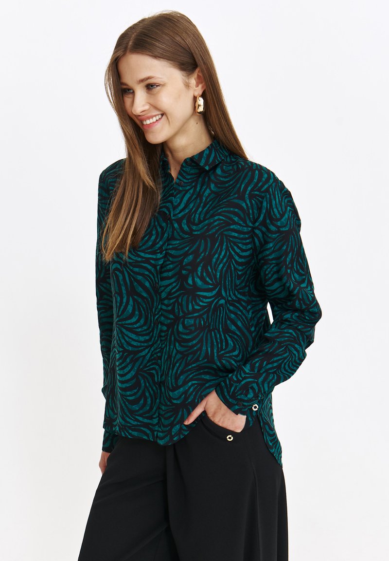 Czarna bluzka z turkusowym, abstrakcyjnym wzorem fali, długie rękawy, kołnierz zapinany na guziki, detal kieszeni oraz lekki połysk. Gładka tekstura materiału.