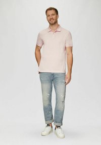 Mann, der lächelt, mit Händen in den Taschen steht, ein hellrosa Poloshirt, ausgewaschene blaue Jeans, die an den Knöcheln hochgerollt sind, und weiße Sneaker trägt.