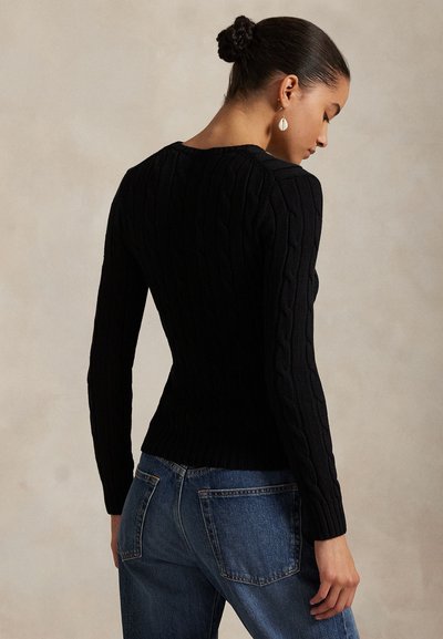 Donna con i capelli raccolti in uno chignon che indossa un maglione nero a trecce e jeans blu, vista di profilo su uno sfondo neutro.