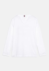 Bílá dlouhá košile s dlouhým rukávem vyrobená z hladké bavlny. Má jemný výstřih do V a barevný akcent na zadním výstřihu s červenými, bílými a modrými pruhy.