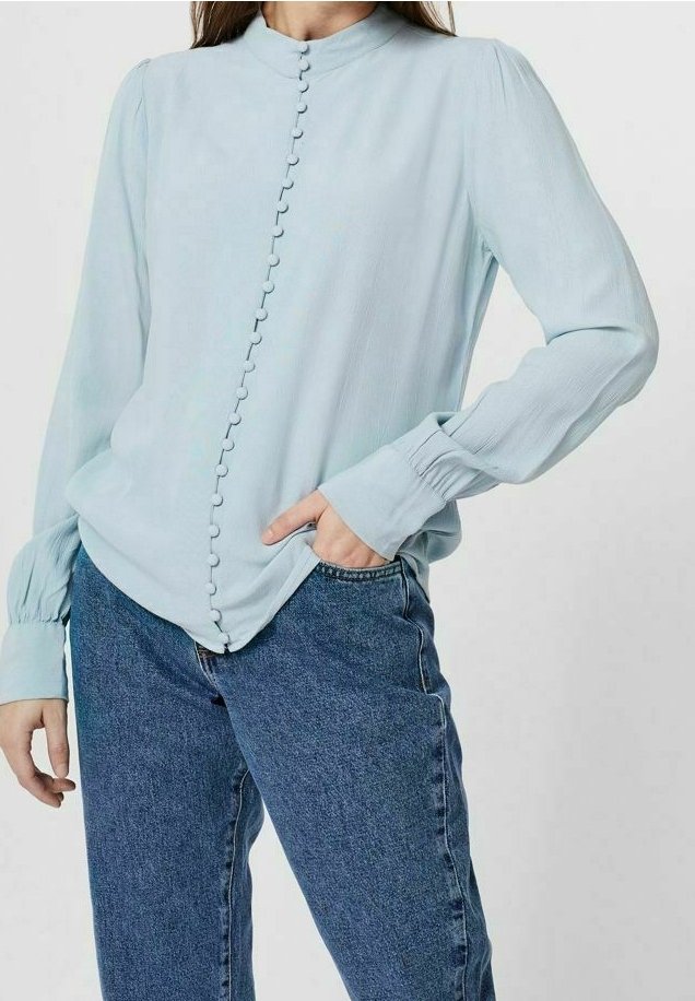 Hellblaue Bluse mit hohem Kragen, Knopfdetails vorne und langen Puffärmeln. Kombiniert mit blauen Jeans. Glatte Textur.