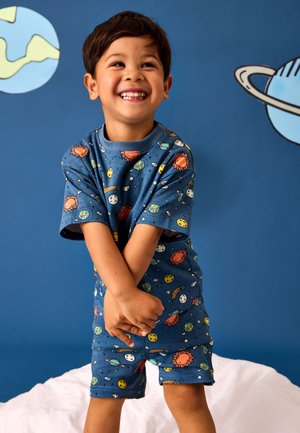 Jeune garçon souriant et croisant les bras, portant un pyjama bleu avec des motifs de soleil, planète et espace sur un fond bleu avec des illustrations de planètes.