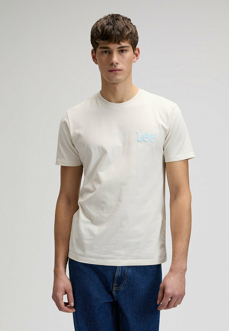 T-shirt crème clair en coton doux, avec un col rond et des manches courtes. Un petit logo "Lee" bleu brodé est situé sur le côté gauche de la poitrine.
