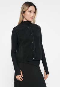 Zwarte fleecedeken met drukknopen, met een textuur en een afgeronde vorm, gedragen over een zwart longsleeve top en jurk.