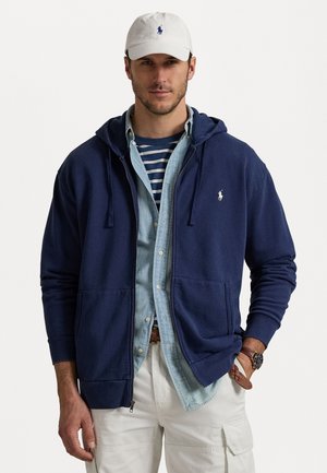 Fato de capuz azul-marinho com fecho éclair e cordões, em material de algodão, com bolsos frontais, logotipo bordado no peito, sobre uma camisa azul-claro.