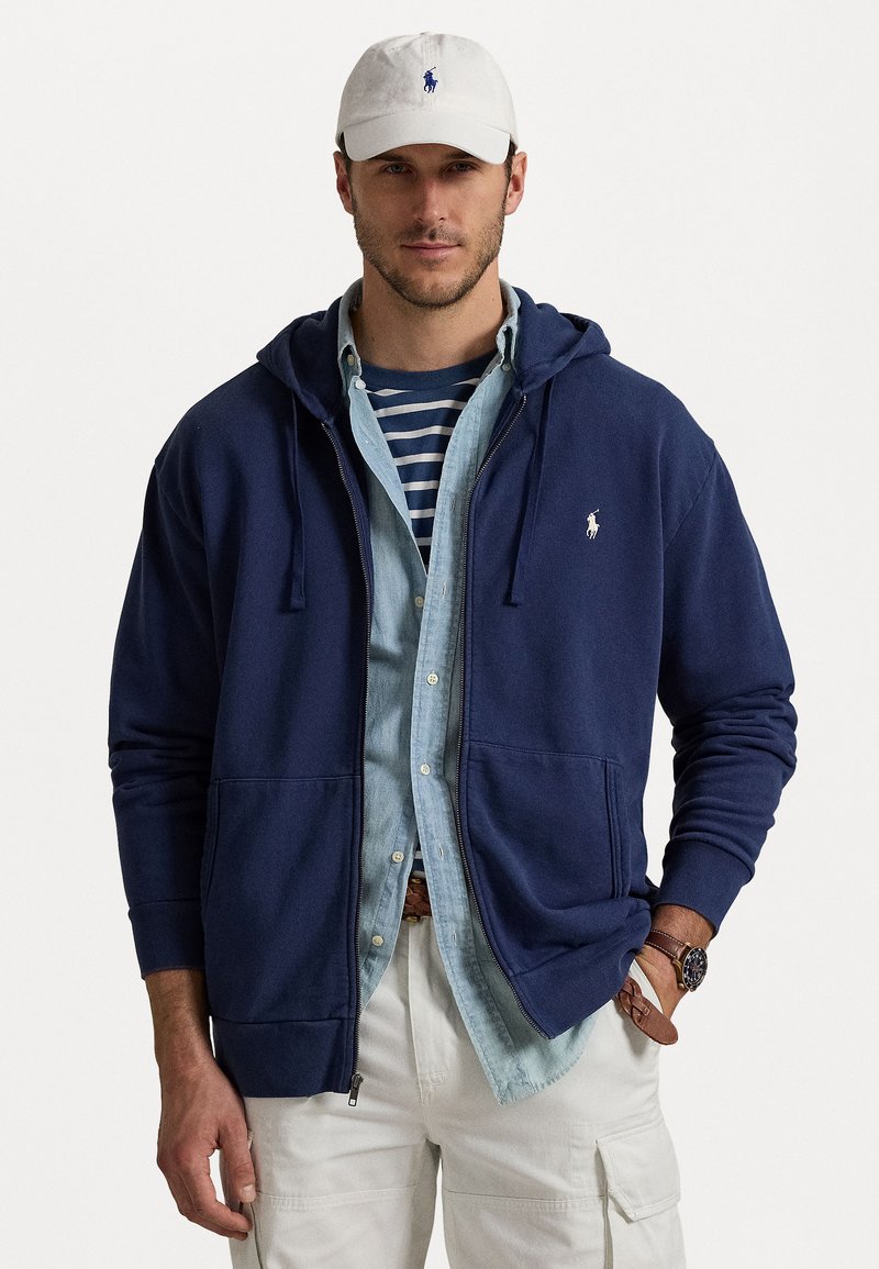 Navyblå zip-up hoodie med dragsko, i bomullsmaterial, framfickor, broderad logga på bröstet, lager på en ljusblå skjorta.