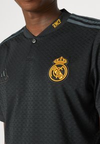 Černý sportovní dres s texturovaným vzorem, zlatě vyšitým klubovým logem, šedými pruhy Adidas a zapínáním na knoflíky s žlutými akcenty.