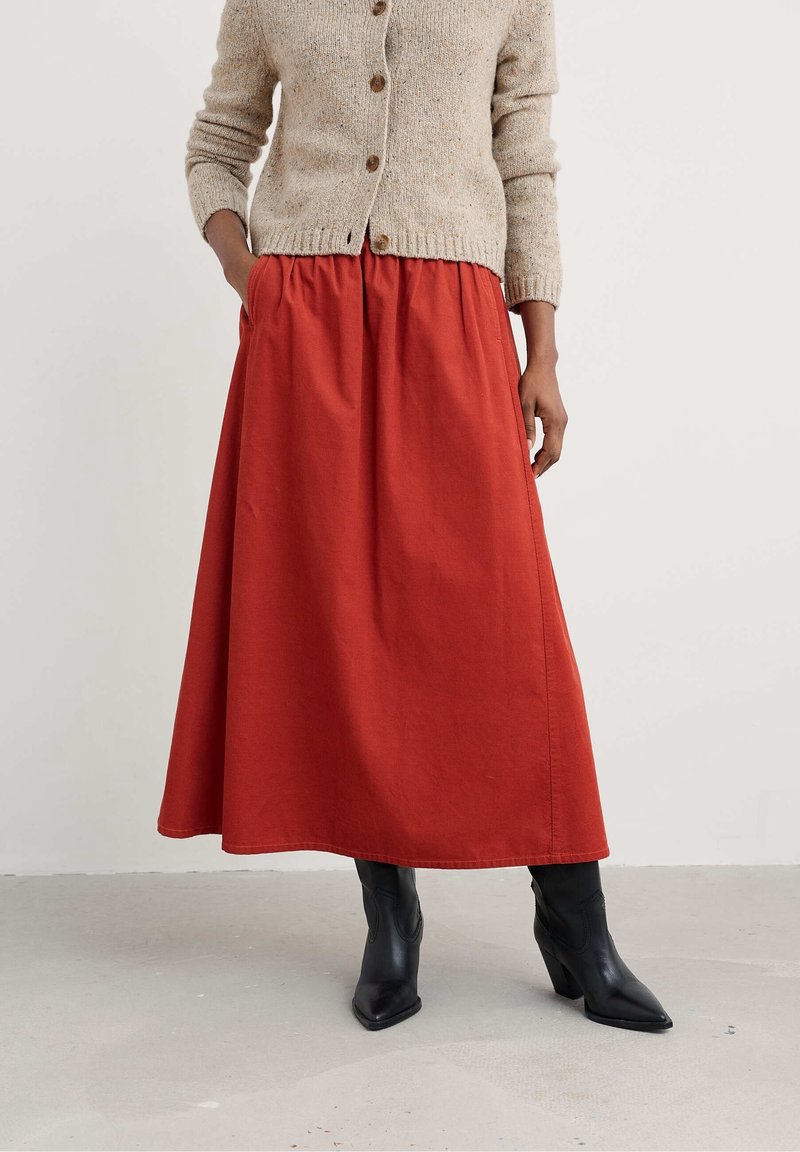 Femme portant un cardigan beige boutonné, une longue jupe rouge avec des poches et des bottines noires à bout pointu, debout sur un sol gris.