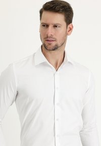 Camisa blanca de manga larga con cuello clásico, cierre de botones y tela suave. Diseño ajustado con un sutil brillo y sin patrones visibles.