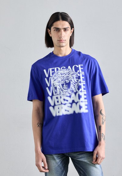 Tricou din bumbac de culoare albastru regal, cu text alb "VERSACE" și un logo proeminent cu Medusa. Mâneci scurte și o croială lejeră.