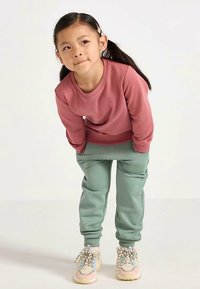 Rosa Sweatshirt mit Rundhalsausschnitt, kombiniert mit hellgrünen Jogginghosen. Das Outfit wird durch pastellfarbene Sneaker mit strukturierten Schnürsenkeln ergänzt.