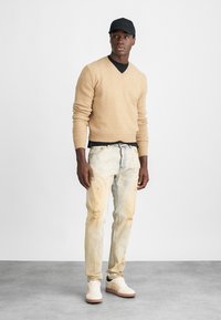 Μπεζ πλεκτό πουλόβερ, μαύρη μπλούζα από κάτω, ανοιχτόχρωμα τζιν ελαφρώς φθαρμένα με ραφές, και λευκά αθλητικά παπούτσια με καφέ σόλες. Μαύρο καπέλο με λογότυπο.