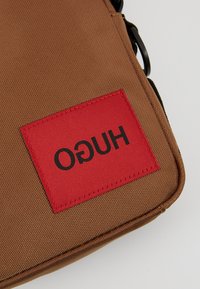 Brązowy, teksturowany materiałowy róg torby z czerwoną prostokątną naszywką, na której jest przeszyty odwrócony czarny napis „HUGO”.