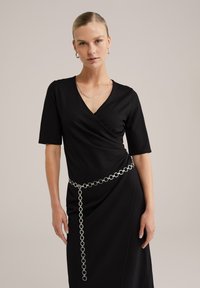 Musta pakettimekko lyhyillä hihoilla, jossa on hopeinen pyöreä ketjupanta. Sujuva pinta ja V-pääntie. Yksinkertainen eleganssi.