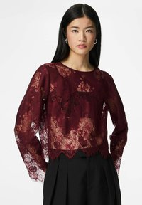 Vīnogkrāsas mežģīņu garroku tops ar ziedu rakstiem un viļņotu apakšu, kārtots pār vienkrāsainu melnu apakšdaļu, demonstrējot caurspīdīgu un teksturētu audumu.