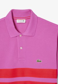 Rosa pikétröja med röda horisontella ränder, ribbad krage, två knappar och en grön krokodil-logotyp. Strukturtyg med en klassisk passform.