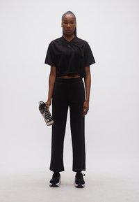 Γυναίκα με μαύρη αθλητική ενδυμασία που κρατάει ένα διαφανές μπουκάλι νερού με μεγάλα μαύρα γράμματα, στέκεται μπροστά σε απλό λευκό φόντο.