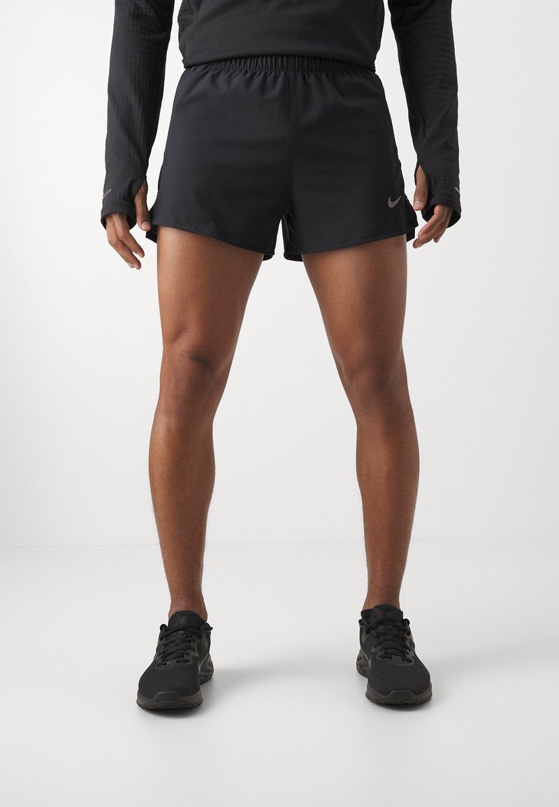 Personne portant un short de sport noir, un haut manches longues noir avec des trous pour les pouces et des chaussures de course noires, debout sur un sol blanc.