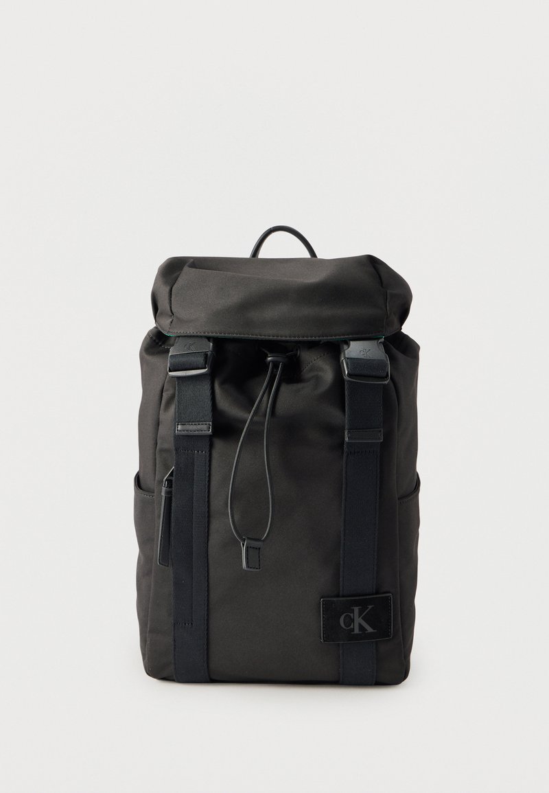 Μαύρο σακίδιο με επάνω καπάκι, σφικτή κλείσιμο, δύο μπροστινές ιμάντες με αγκράφες, πλαϊνές τσέπες και πατς με το λογότυπο Calvin Klein στο εμπρόσθιο κάτω δεξί μέρος.