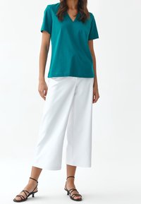 Teal bavlněné tričko s výstřihem do V, spárované s bílými širokými kalhotami a černými sandály na řemínky. Jednoduchý design, univerzální barvy.