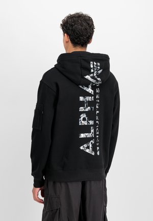 Sudadera negra con una gran gráfica reflectante blanca que dice "ALPHA" verticalmente en la parte posterior. Cuenta con un bolsillo tipo canguro y una tela texturizada.