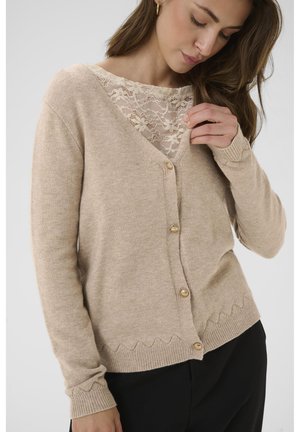 Beige Strickjacke aus weichem Material, mit V-Ausschnitt, gewelltem Saum und goldenen Knöpfen. Über einem Spitzenshirt getragen.