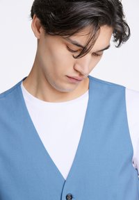 Ljusblå, ärmlös väst med en slät textur, med v-ringning och knappstängning fram, lager över en vit t-shirt.