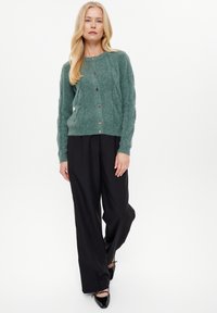 Grüner Strickcardigan mit Rundhalsausschnitt, Knopfverschluss und strukturiertem Muster, kombiniert mit schwarzen weiten Hosen und schwarzen Absatzschuhen.