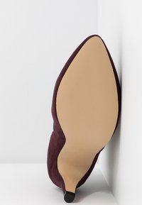 Scarpa con tacco alto in suede bordeaux, con punta affilata e suola beige liscia, caratterizzata da un design elegante e minimalista e dettagli testurizzati.