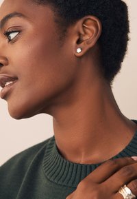 Silver stud earring, round and shiny; model wears a dark green ribbed sweater. Close-up of neck and ear, showing the earring clearly.