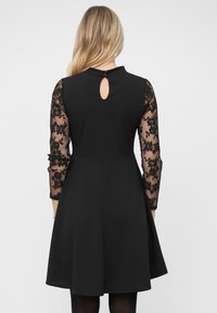 Vestido negro con mangas de encaje translúcido hasta el codo que presentan patrones florales. Un detalle en forma de ojo de cerradura se encuentra en la parte posterior del escote.