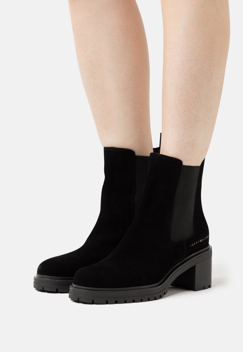 Bottes à cheville en daim noir avec talons blocs et panneaux élastiques sur les côtés, portées sur jambes nues, sur fond blanc.