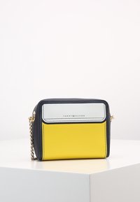 Kompaktowa torba w kolorach granatowym, żółtym i białym, z łańcuszkowym paskiem, gładką fakturą oraz logo Tommy Hilfiger z przodu.