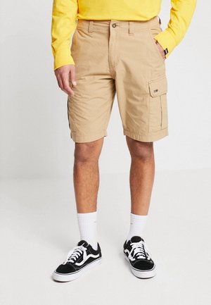Kakifarvede cargoshorts med sidfickor, tillverkade av bomullsblandat tyg. Stylad med en gul långärmad topp och svarta sneakers med vita detaljer.
