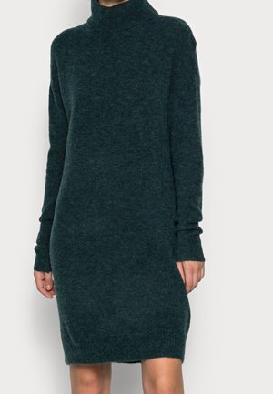 Tmavě zelené šaty s rolákem vyrobené z měkké, strukturované pleteniny. Mají dlouhé rukávy a volný střih se mírně zakřiveným lemem.