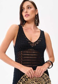 Schwarzes gestricktes Kleid mit einem transparenten Design, das große offene Webmuster aufweist. Das Modell hält einen Strohhut und ist mit Ohrringen und einem Armreif accessoriert.