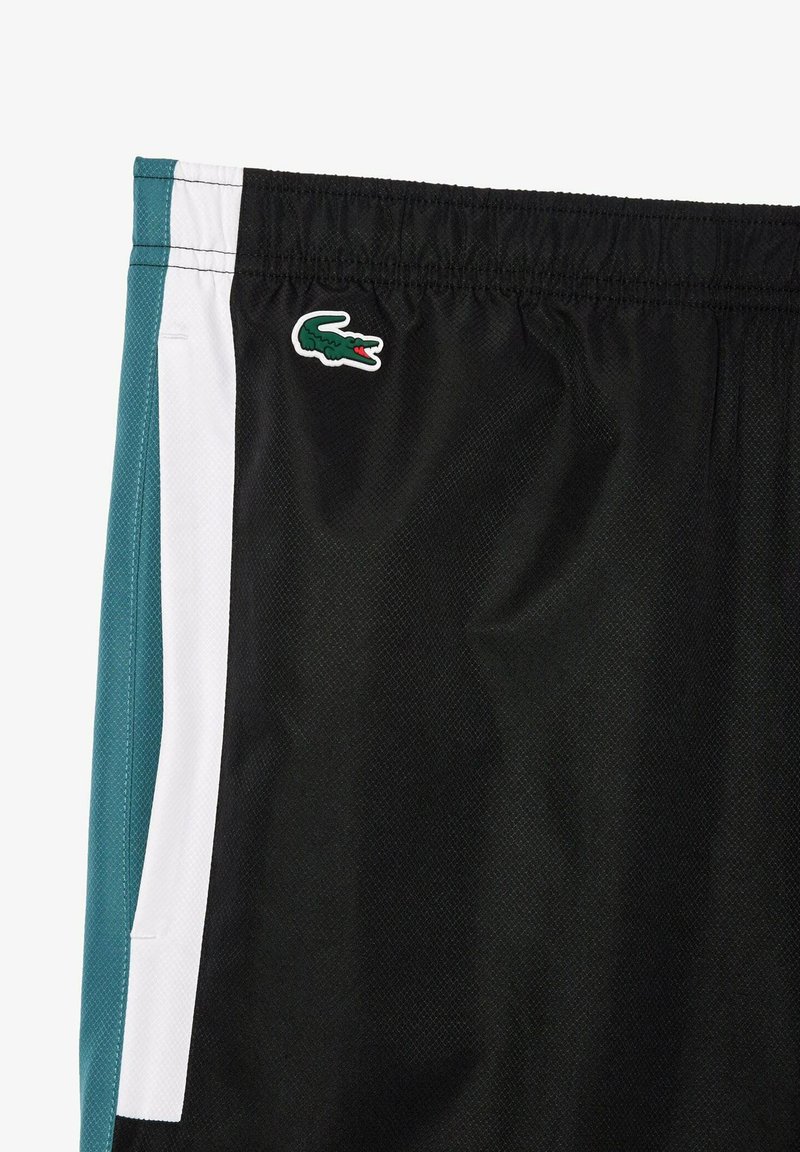 Shorts de sport noirs avec un tissu texturé, des panneaux latéraux blancs et bleu canard, et un logo de crocodile vert sur la cuisse gauche.