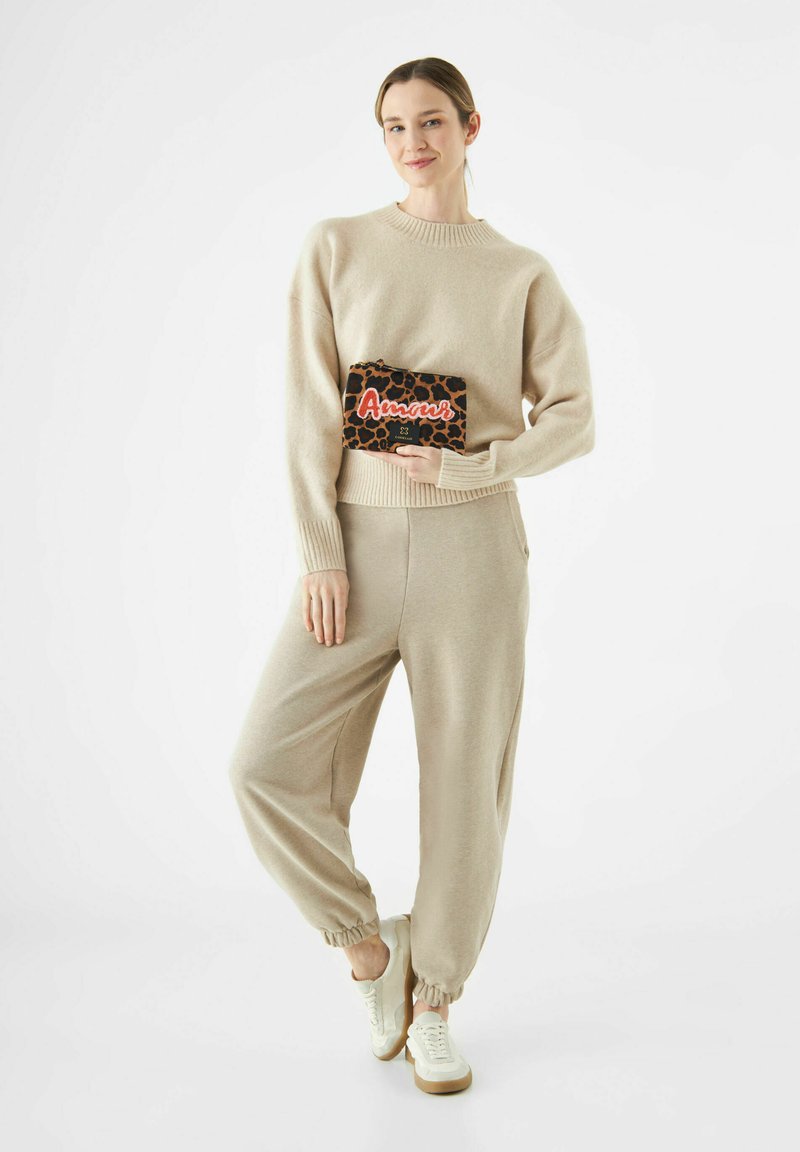 Vrouw in een beige trui en joggers, met een luipaardprint clutch met de tekst "Amour", staand met één been gekruist en glimlachend.