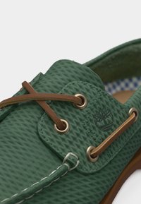 Πράσινο δερμάτινο παπούτσι με υφή, καφέ δερμάτινα κορδόνια περασμένα από μεταλλικά τεφτέρια και ραμμένες λεπτομέρειες κοντά στο σήμα.