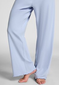 Blote voeten met lichtblauwe, geribbelde, wijde broek met gekrulde randen, tegen een neutrale achtergrond.